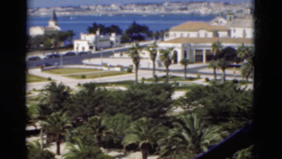 1975 Portugal 8mm frame showing a white waterfront building with terracotta roof, palms, gardens, road, and distant shoreline across the bay.