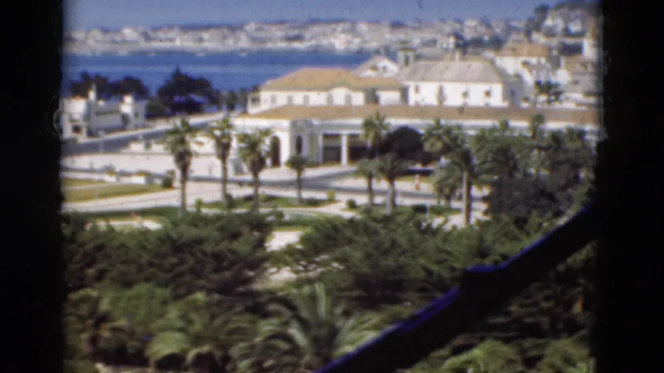 Final 1975 Portugal view of palms and a white coastal building beside the road, with blue bay water and a distant city skyline.