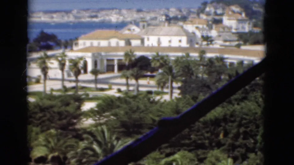 1975 Portugal home-movie view of white coastal buildings, palms, greenery, and sea seen through a window frame.