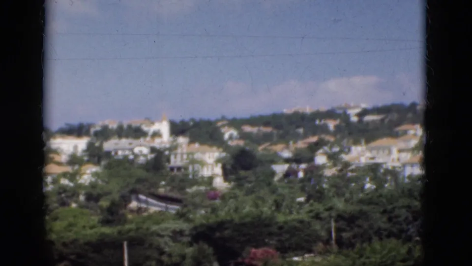 1975 Portugal hillside townscape with white buildings, terracotta roofs, trees, and a dark vertical window bar.