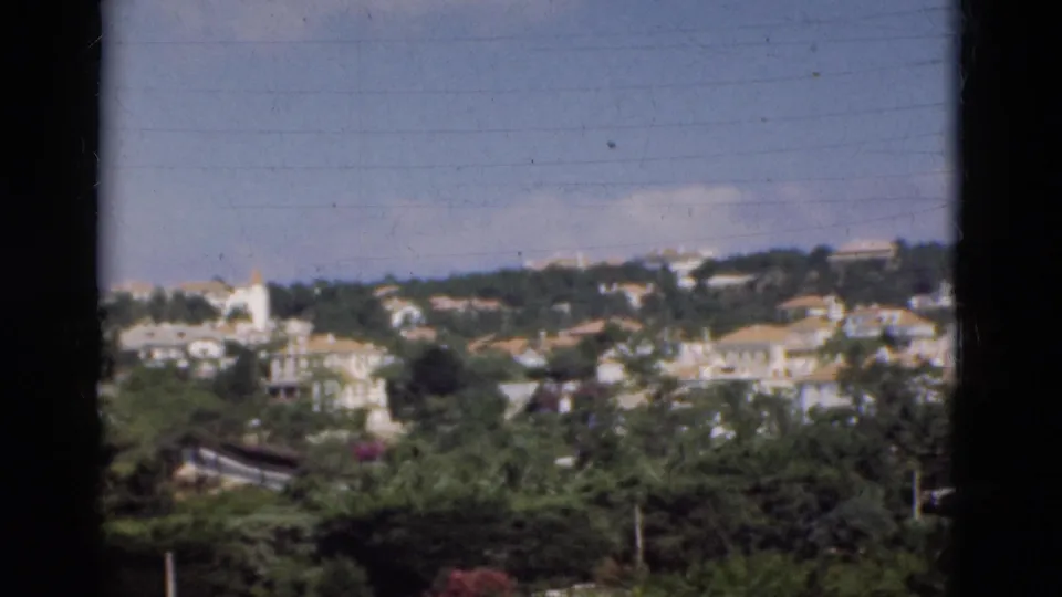 1975 Portugal home-movie frame showing a leafy hillside, white houses, pale roofs, and distant ridge buildings.