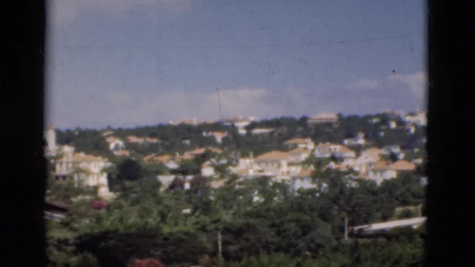 1975 Portugal view across dense trees toward hillside houses and pale-roofed buildings under blue sky.