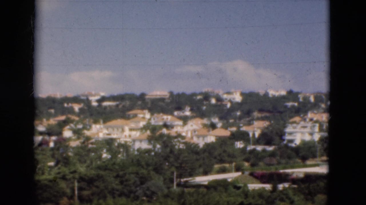 Hillside town with pale houses, trees, and a road in the foreground.