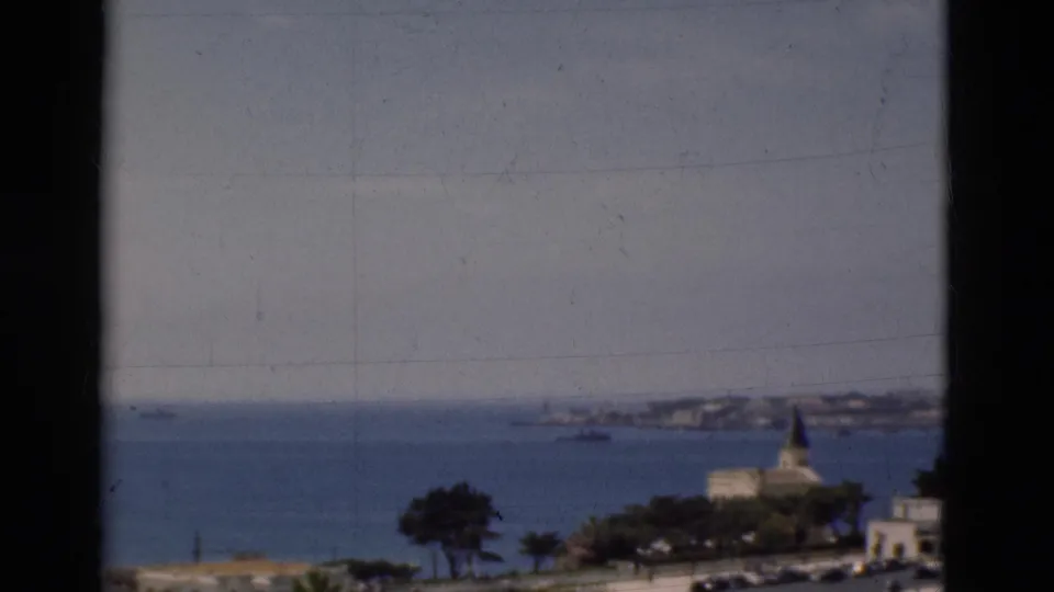 Soft 1975 Portugal home-movie frame of the coastal water, distant land, small boats, trees, and a church steeple.