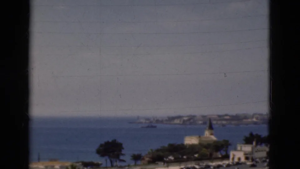 1975 Portugal 8mm view of a blue bay, church spire, distant shoreline, and hazy sky seen through dark edge framing.