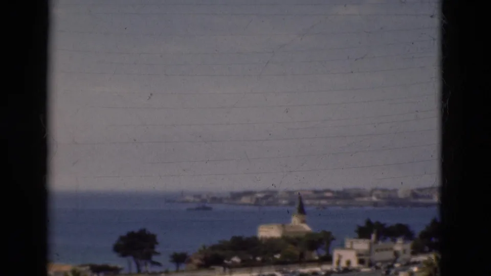 1975 Portugal home-movie frame showing the bay, church spire, distant town shoreline, trees, and dark vertical framing.