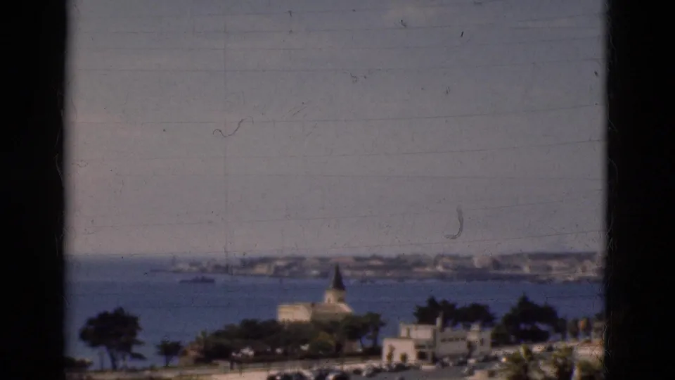Soft 8mm frame from Portugal in 1975 showing a bay, church spire, distant shoreline, trees, and broad hazy sky.