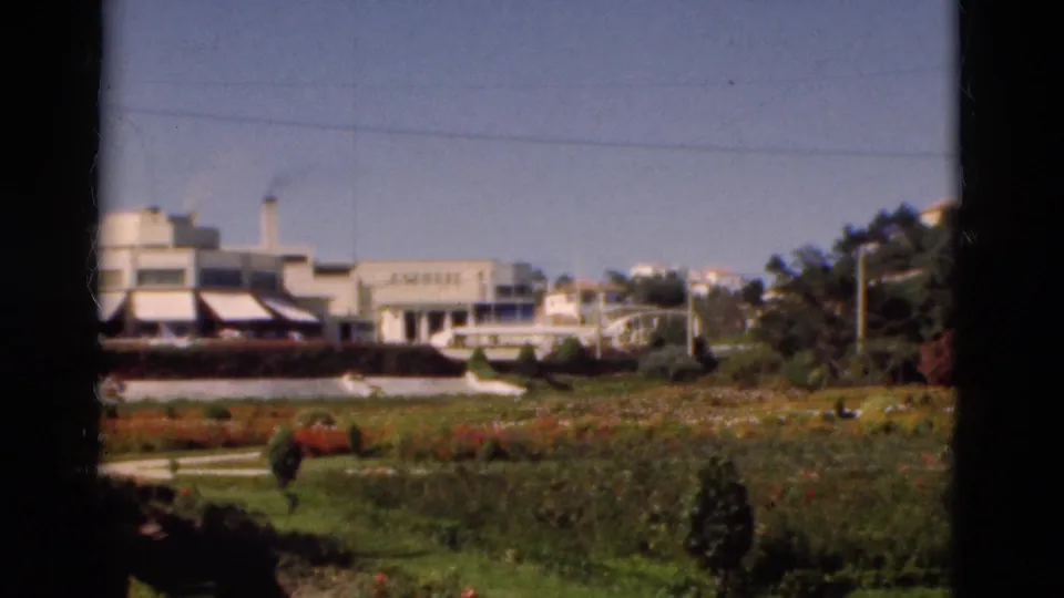 Soft 8mm Portugal frame with a white building, low awnings, chimney, garden beds, shrubs, and dark framing from the camera view.