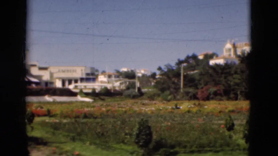 1975 Portugal 8mm frame showing garden beds, trees, white buildings, and a distant domed structure under a blue sky.