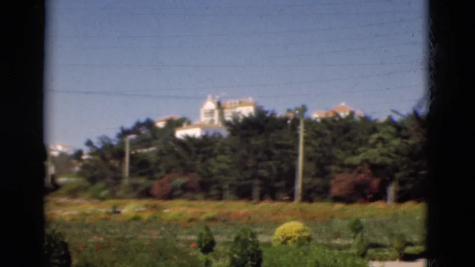 1975 Portugal 8mm frame with planted lawns, low flowers, trees, utility poles, and distant white buildings seen through foreground bars.