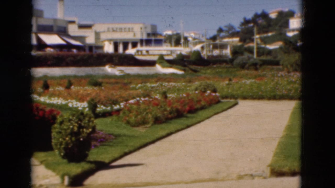 1975 Portugal 8mm frame showing a pale garden path, colorful flowerbeds, lawns, buildings, and moored boats beyond.