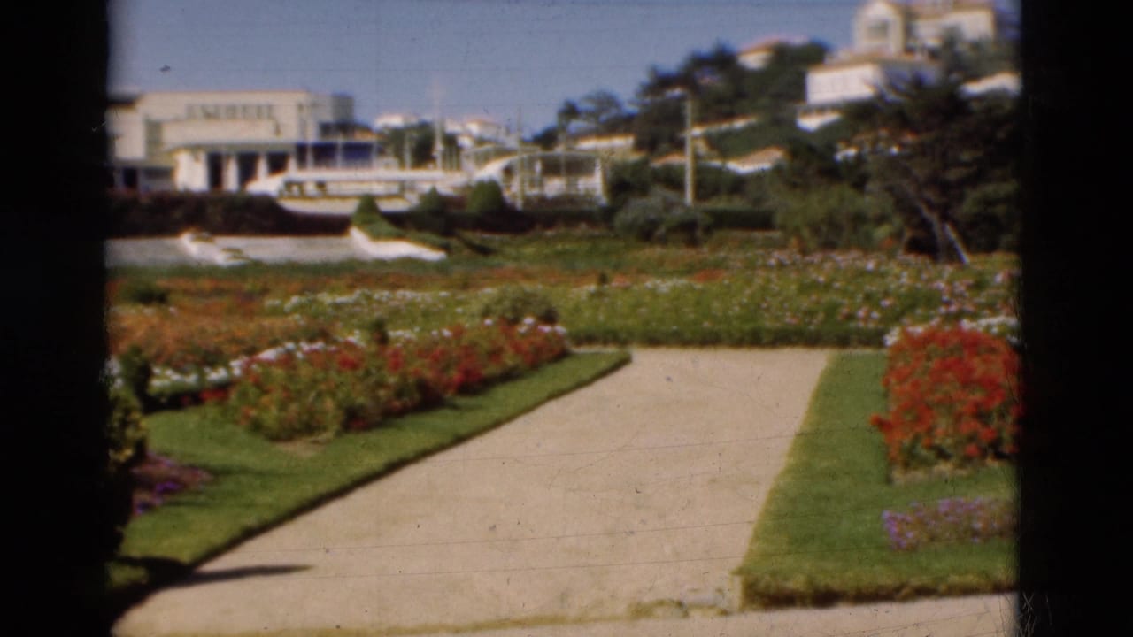 1975 Portugal 8mm frame with a walkway, red and white flowers, manicured grass, boats, and hillside buildings under blue sky.