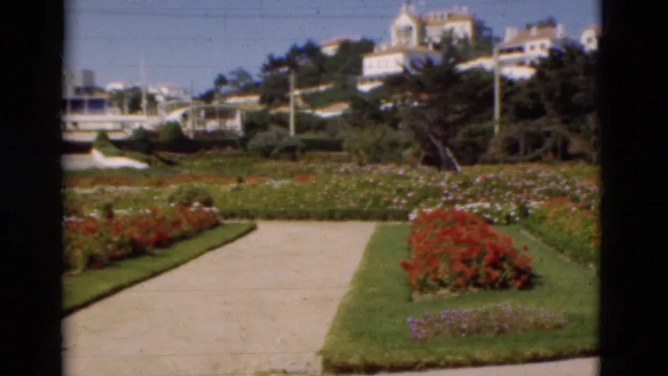1975 Portugal 8mm frame showing the path edge, flowerbeds, green lawn, moored boats, trees, and white hillside houses.