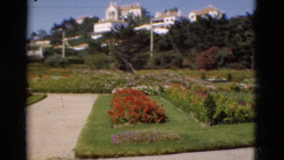 1975 Portugal 8mm frame with red blooms, a pale path, manicured lawn, dense trees, and white buildings on the hillside.