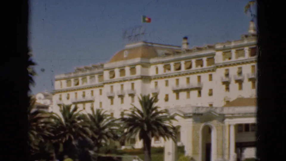 1975 Portugal 8mm frame of a white official-looking building with palm trees and a Portuguese flag flying above the roofline.