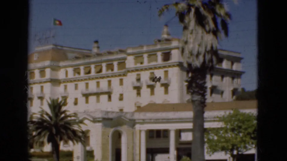 1975 Portugal 8mm frame of the official-looking white building, rooftop flag, palm trees, and greenery in the foreground.