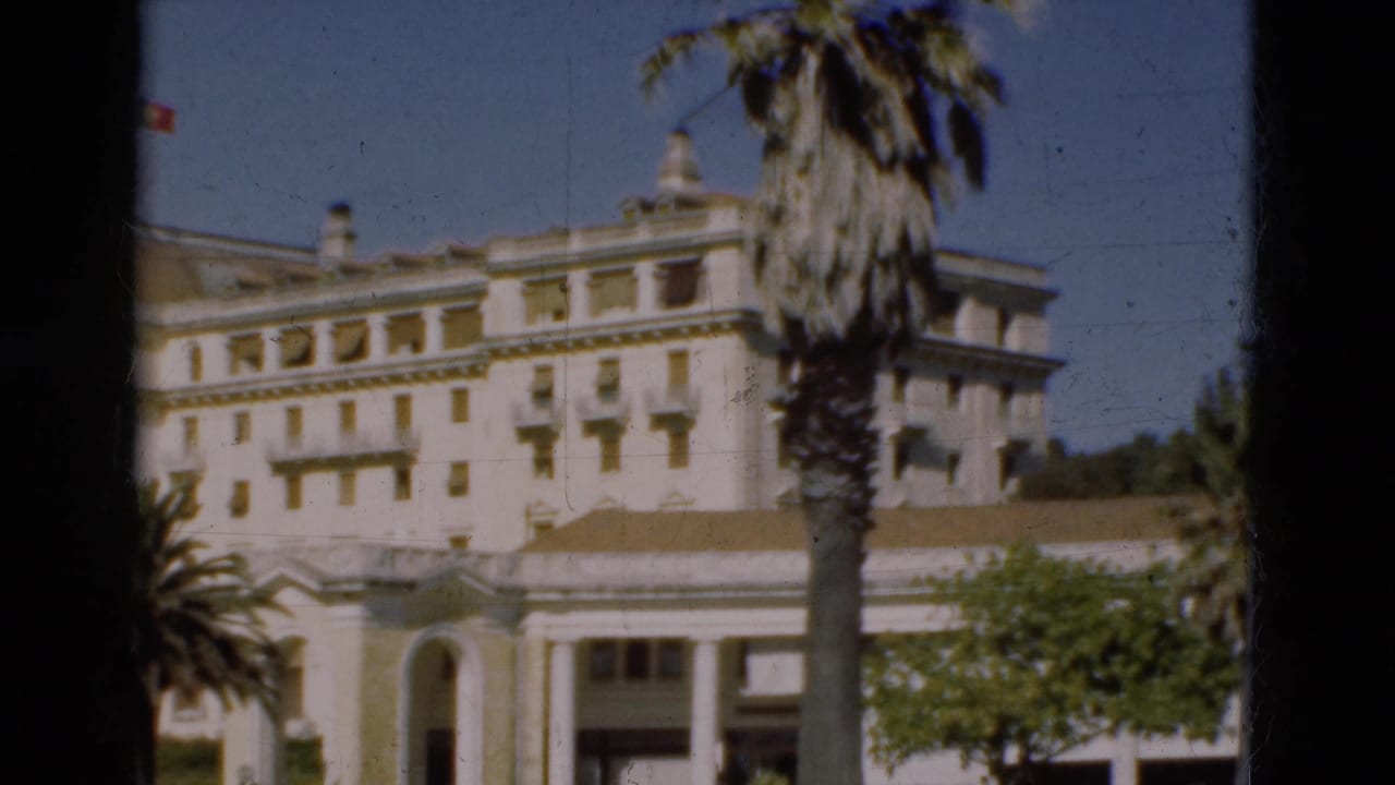 1975 Portugal 8mm frame showing a white classical building with balconies, arched entrance, palm trees, and blue sky.