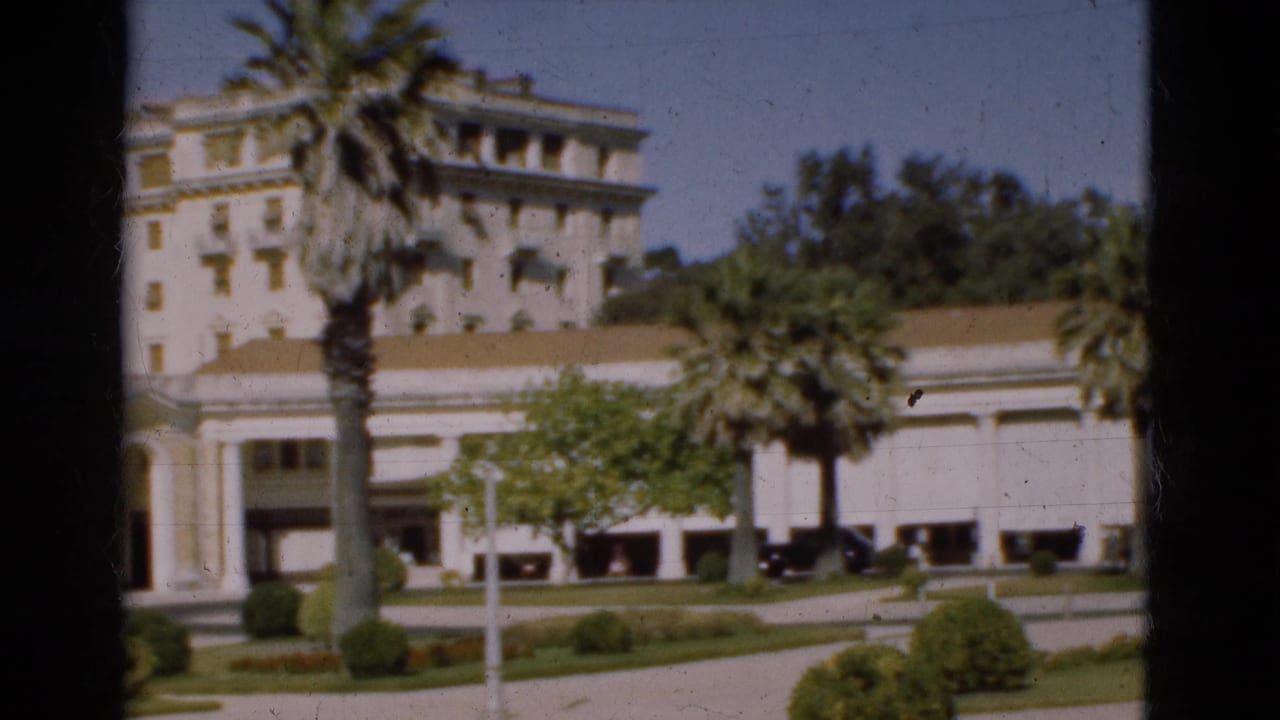 1975 Portugal 8mm frame of a formal white facade with columns, balconies, palm trees, trimmed shrubs, and a driveway.