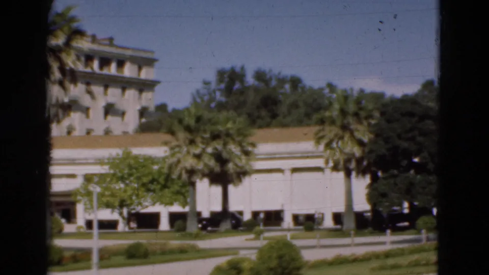 1975 Portugal 8mm frame showing part of the white building, palm trees, low landscaping, driveway, and parked cars.