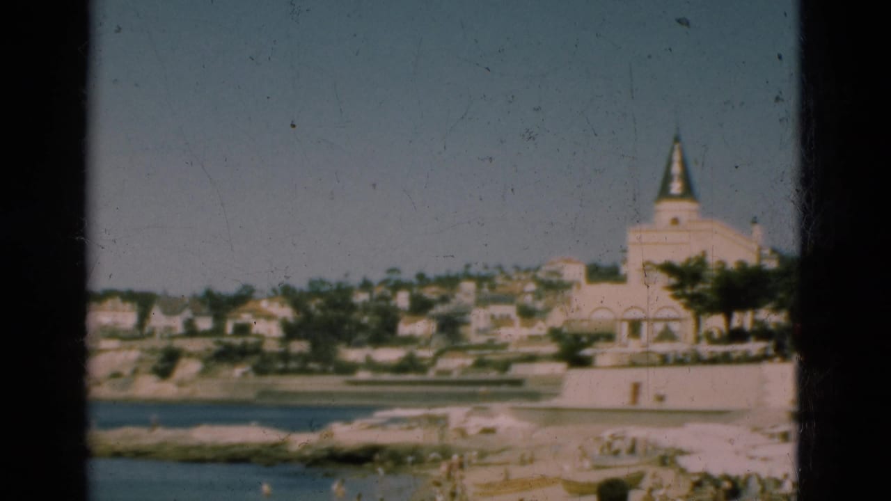 1975 Portugal 8mm frame showing a coastal town across the water with a large pale spired building and terraced houses.
