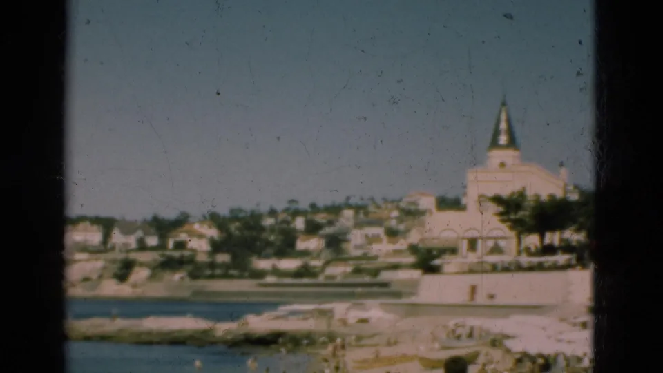 1975 Portugal 8mm frame showing a coastal town across the water with a large pale spired building and terraced houses.