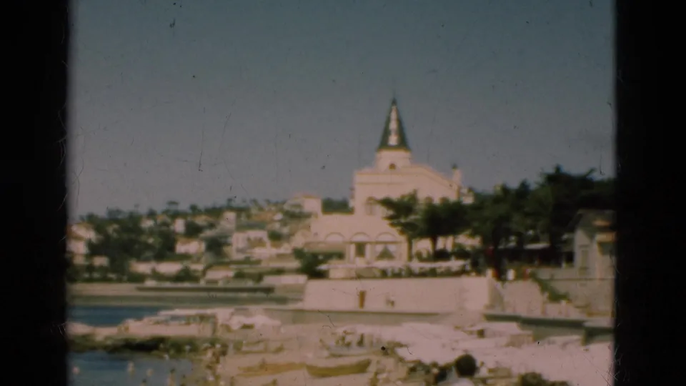 1975 Portugal waterfront view with a spired pale building, trees, low hillside homes, and beach rocks in the foreground.
