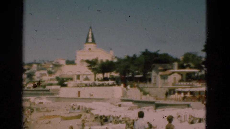 1975 Portugal 8mm frame of a beach and waterfront, with people, small boats, trees, and a spired building in the background.