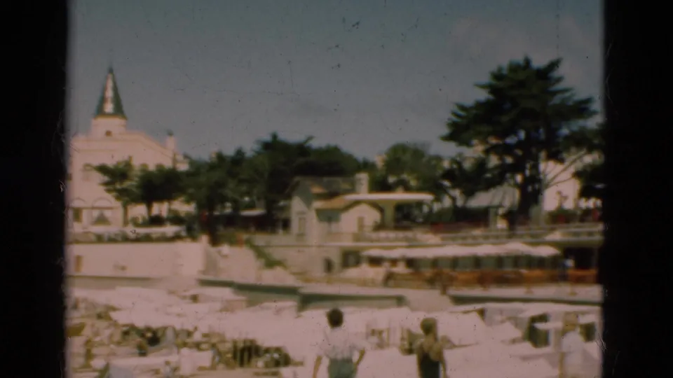 1975 Portugal coastal frame showing beachgoers and boats below waterfront buildings, trees, and a partly obscured spired structure.