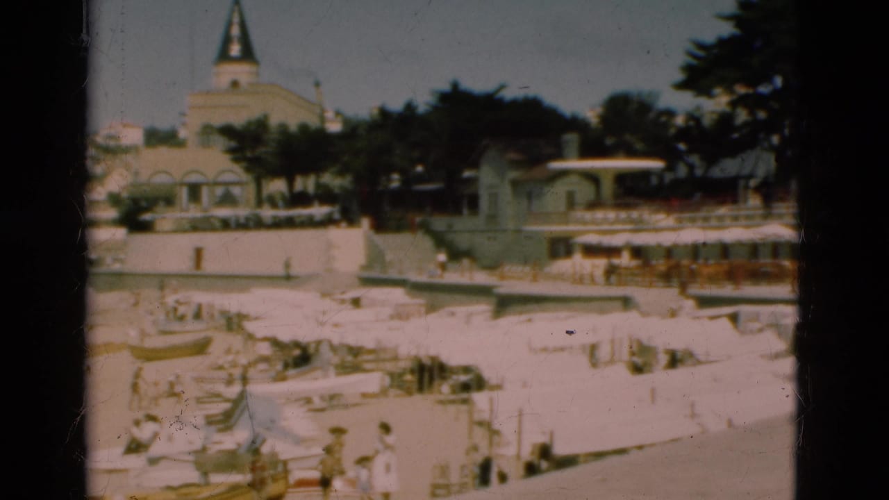 Beachfront with white umbrellas, distant people, promenade buildings, trees, and a steepled structure.