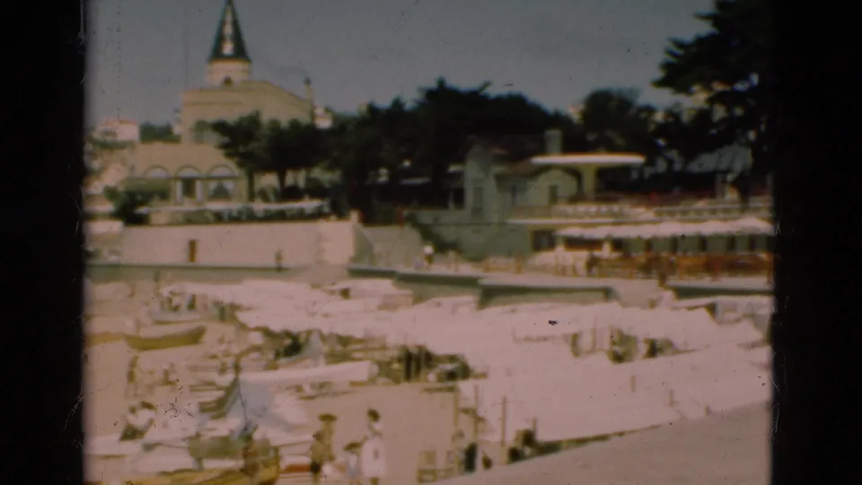 1975 Portugal 8mm frame showing a bright beachfront with white umbrellas, small figures, promenade walls, trees, and a steepled building beyond.
