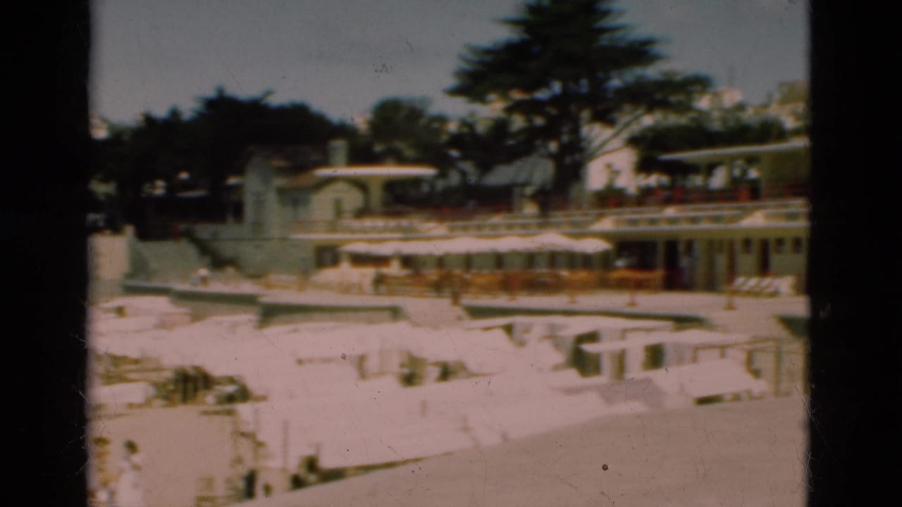 1975 Portugal home-movie frame with beach umbrellas and loungers below promenade buildings, trees, and a steepled structure in the background.