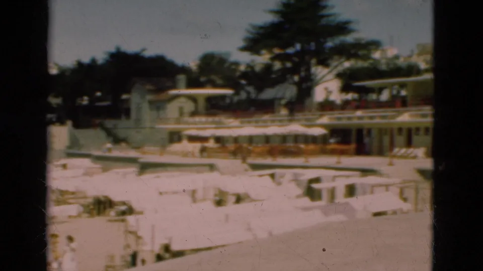 1975 Portugal home-movie frame with beach umbrellas and loungers below promenade buildings, trees, and a steepled structure in the background.