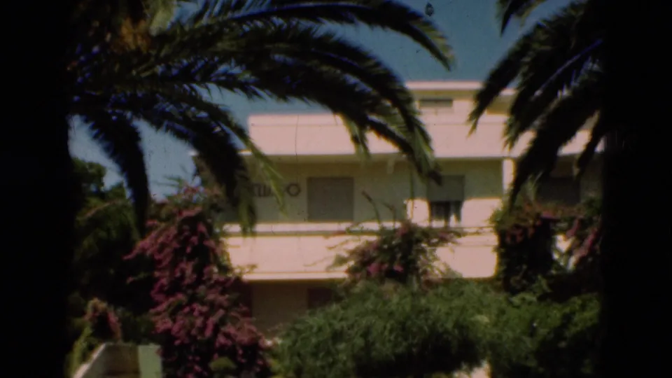1975 Portugal 8mm frame showing palm trees and flowering shrubs before a low white resort building with balconies and windows.
