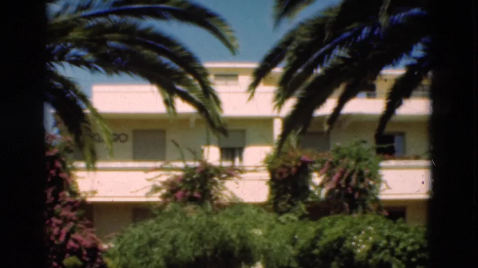 1975 Portugal home-movie frame of a white hotel-like building with balconies, seen through palm fronds and dense coastal landscaping.