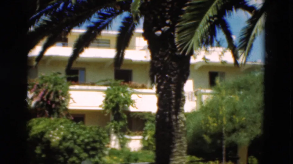 1975 Portugal 8mm still showing a palm trunk in front of a sunlit white building, balcony railings, shrubs, and garden foliage.