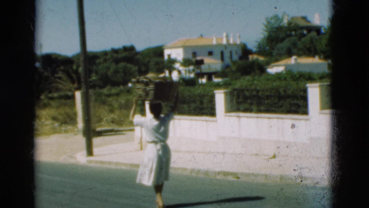 In Portugal 1975 home-movie footage, a woman in white carries a basket on her head beside a white wall and roadside greenery.