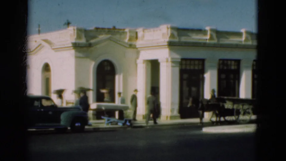 A white Portuguese street building appears with several pedestrians, a parked older car, and a horse-drawn cart in 1975 8mm footage.