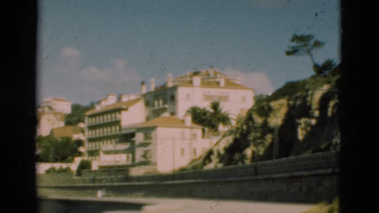 1975 Portugal 8mm frame with white and pale buildings rising on a hillside above a stone wall and palm-lined road edge.