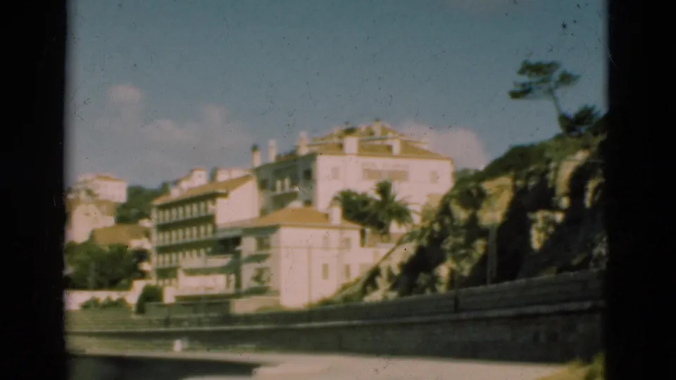 1975 Portugal 8mm frame with white and pale buildings rising on a hillside above a stone wall and palm-lined road edge.
