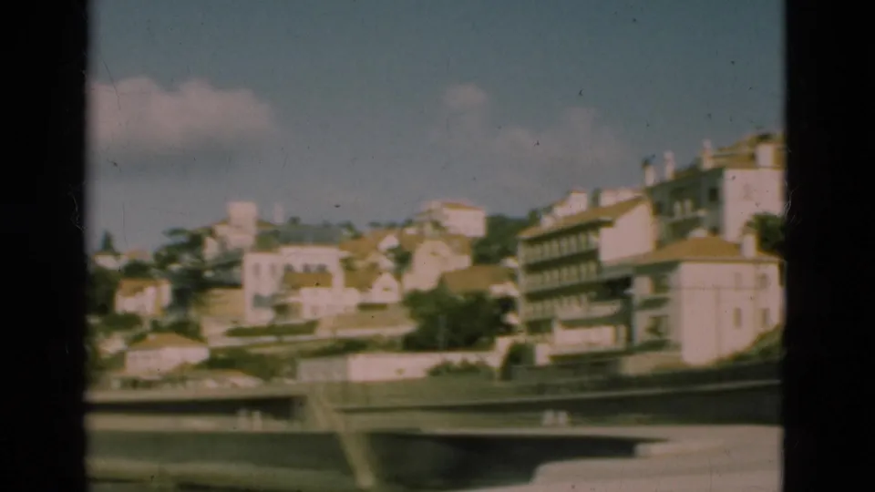 1975 Portugal 8mm frame showing a wider hillside town view with pale buildings, palm trees, masonry walls, and bright sky.