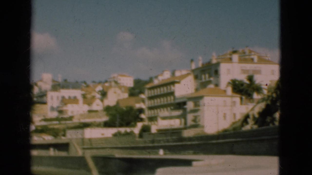 1975 Portugal home-movie frame showing pale hillside buildings above a waterfront wall, seen through dark film borders.