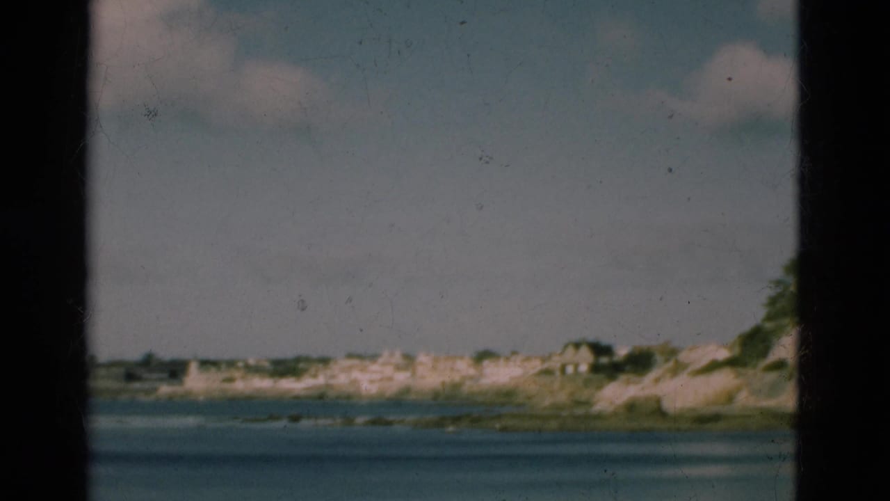 1975 Portugal 8mm frame of calm ocean water and a distant shoreline with light-colored buildings under a pale sky.