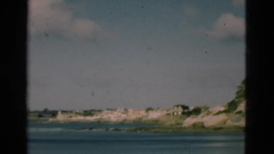 1975 Portugal 8mm frame of calm ocean water and a distant shoreline with light-colored buildings under a pale sky.