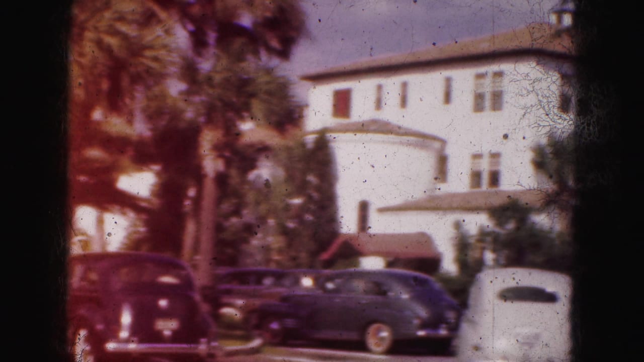 1975 Florida home-movie frame with parked rounded sedans, palm trees, and a large white building in bright daylight.
