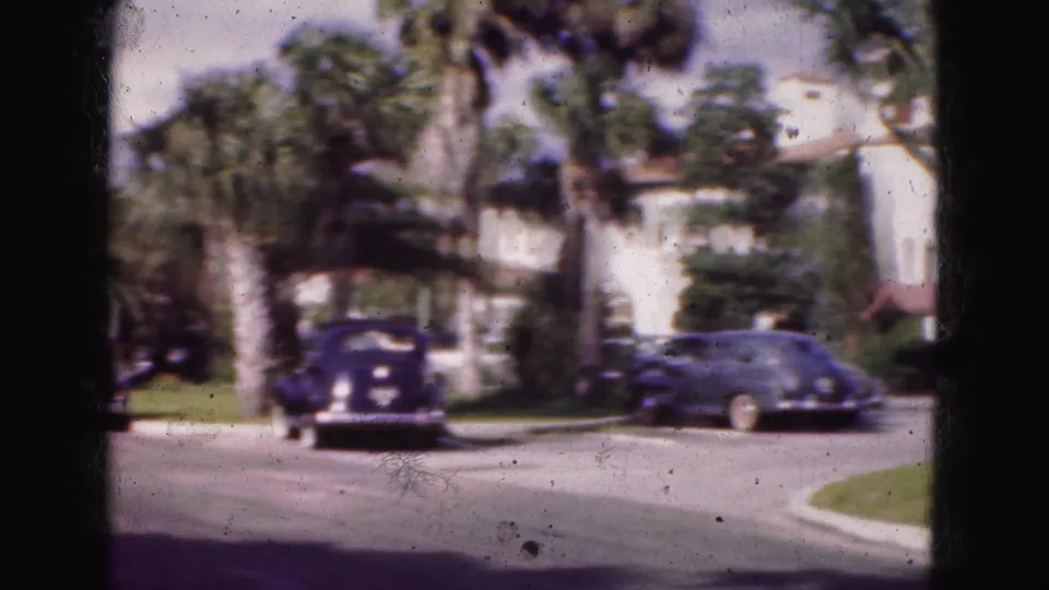 1975 8mm Florida frame showing a residential street with older parked cars, palm trees, lawns, and house fronts.