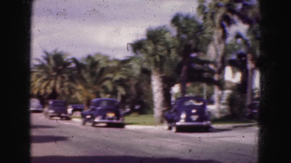 1975 Florida home-movie frame showing a palm-lined suburban street with multiple dark classic cars and low buildings beyond.