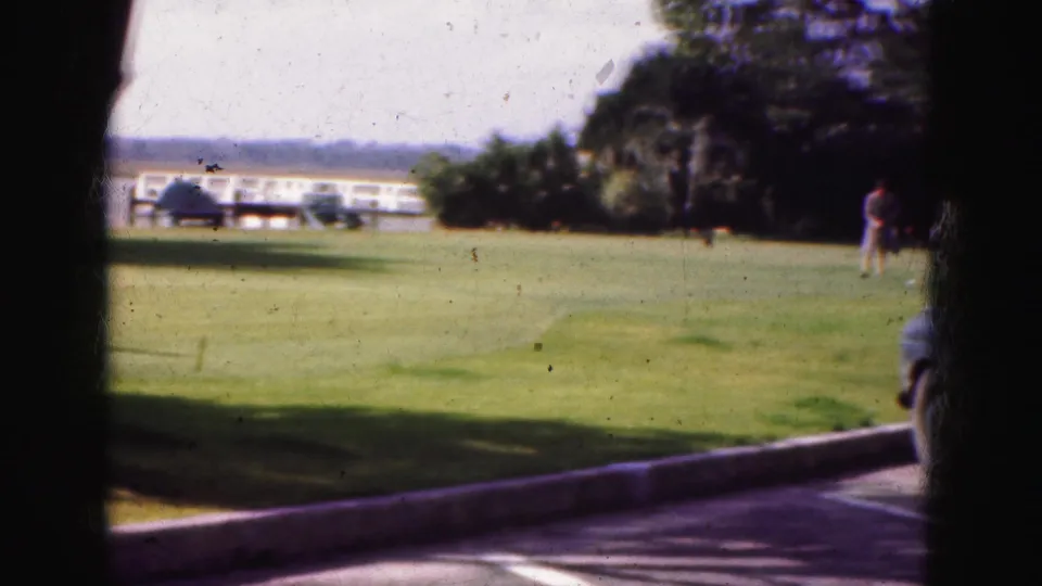 A soft 8mm view of a wide Florida lawn shows a long low building, distant people, trees, and a curb at the road edge.