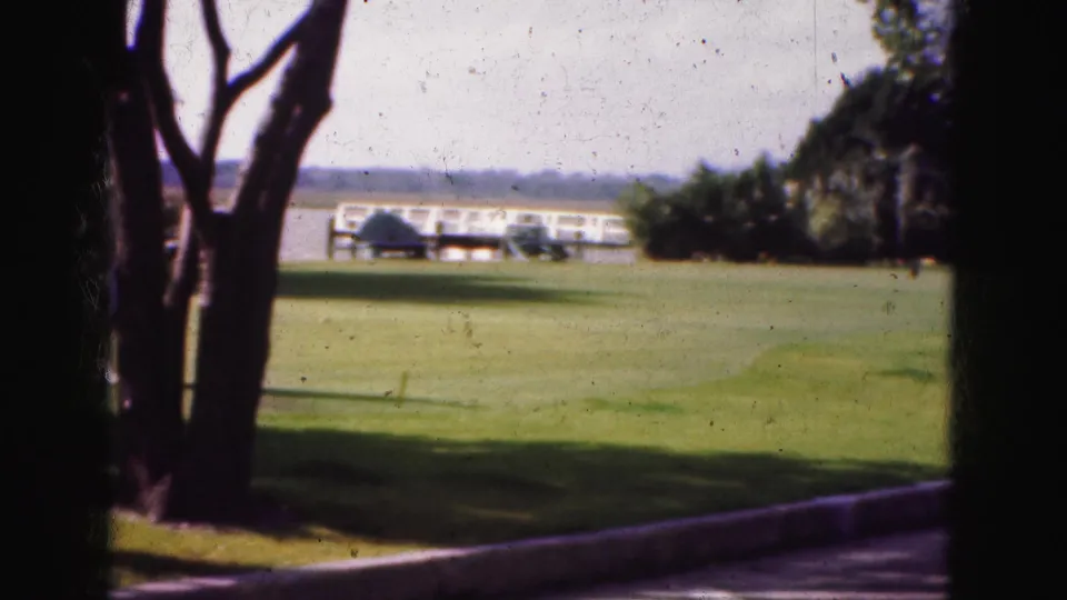 1975 Florida home-movie frame showing a green lawn, tree trunks, and a distant low building beyond landscaped grounds.