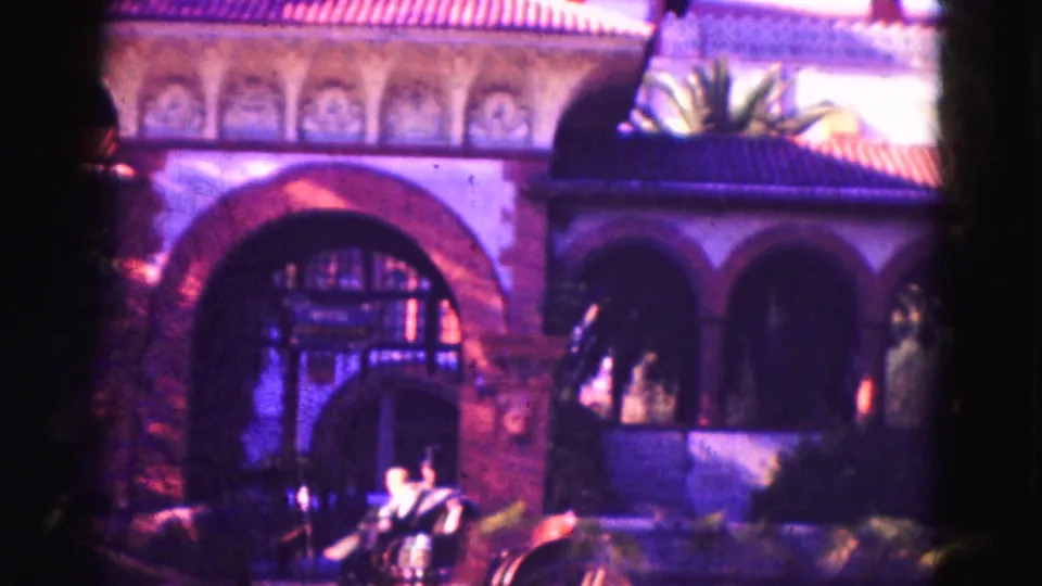 1975 Florida home-movie frame showing the arched facade, decorative trim, palm leaves, and part of the horse-drawn carriage in front.