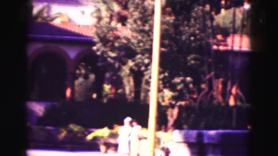1975 Florida home-movie frame showing visitors near a low wall, trees, a pole, and part of the landscaped building grounds.